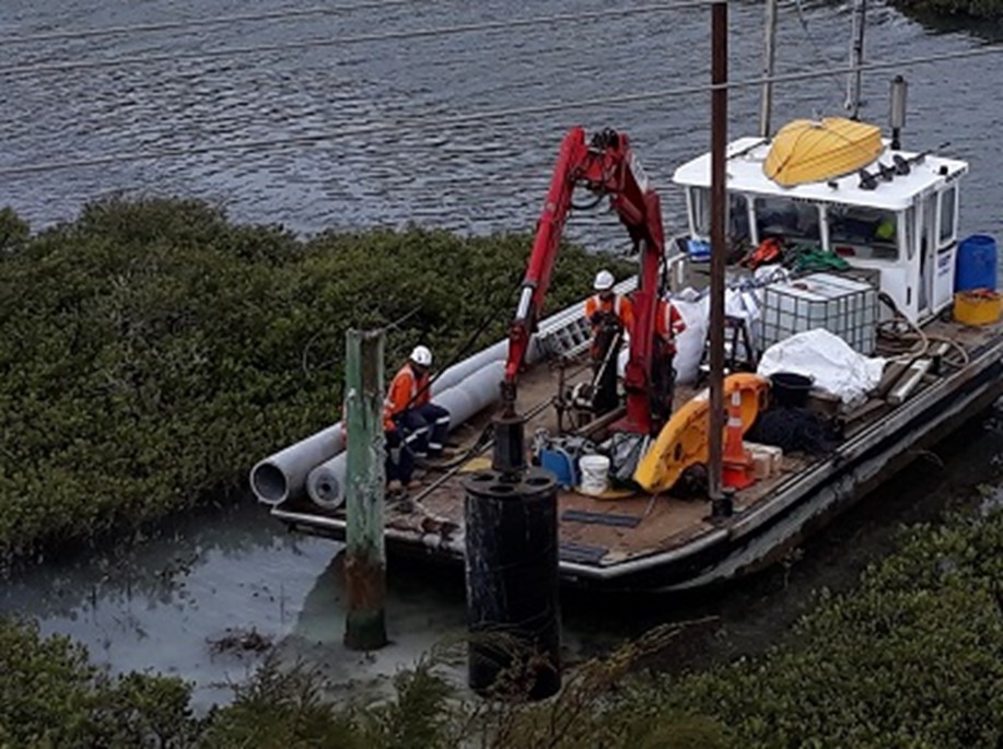 A barge on the side of an estuary with a digger on it and workmen erecting a power poles