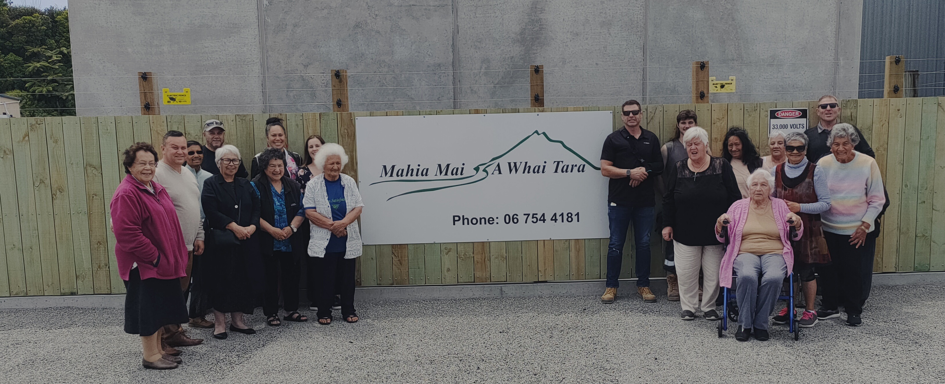 A group of elderly people standing in front of a fence with a sign on it.