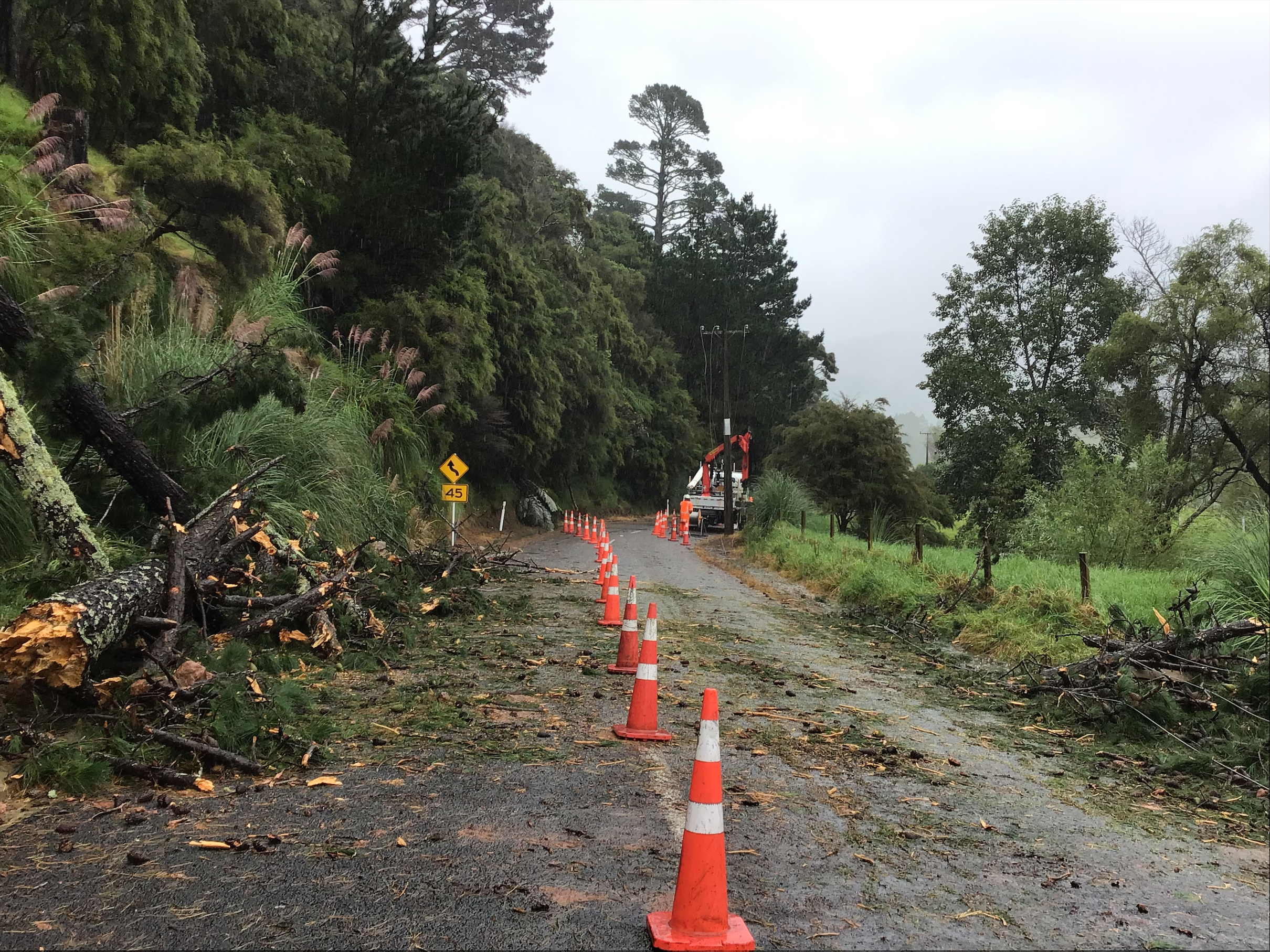 A rural road strewn with tree debris, with orange cones up the centre and a electricity truck in the distance.